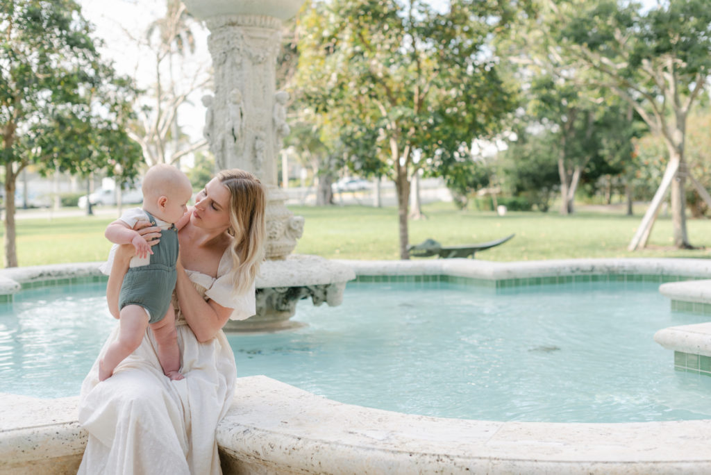 Mom holding up baby looking at baby sitting by the fountain