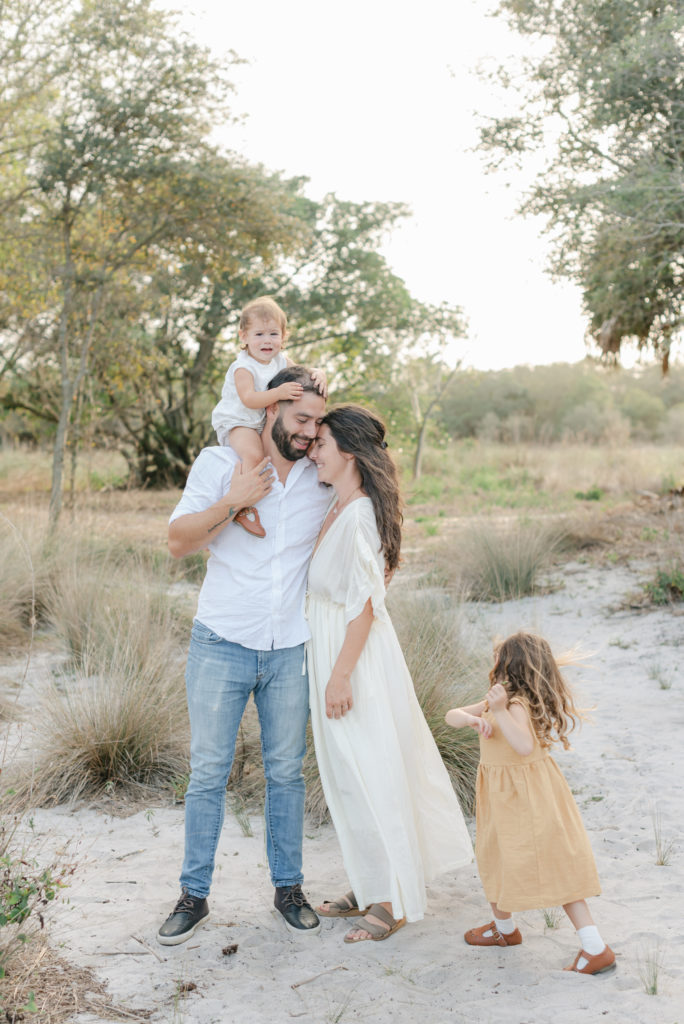 Mom and dad snuggled, daughter on dad's shoulders, other daughter playing