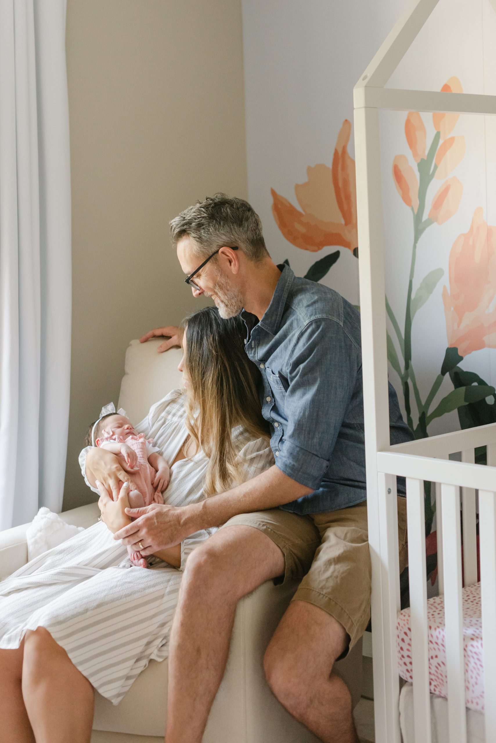 Mom and dad looking down at baby girl, sitting on chair