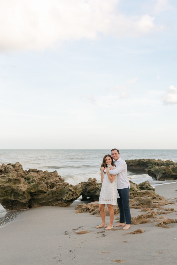 Man wrapping arms around woman in front of rocks on the beach by Jupiter photographer