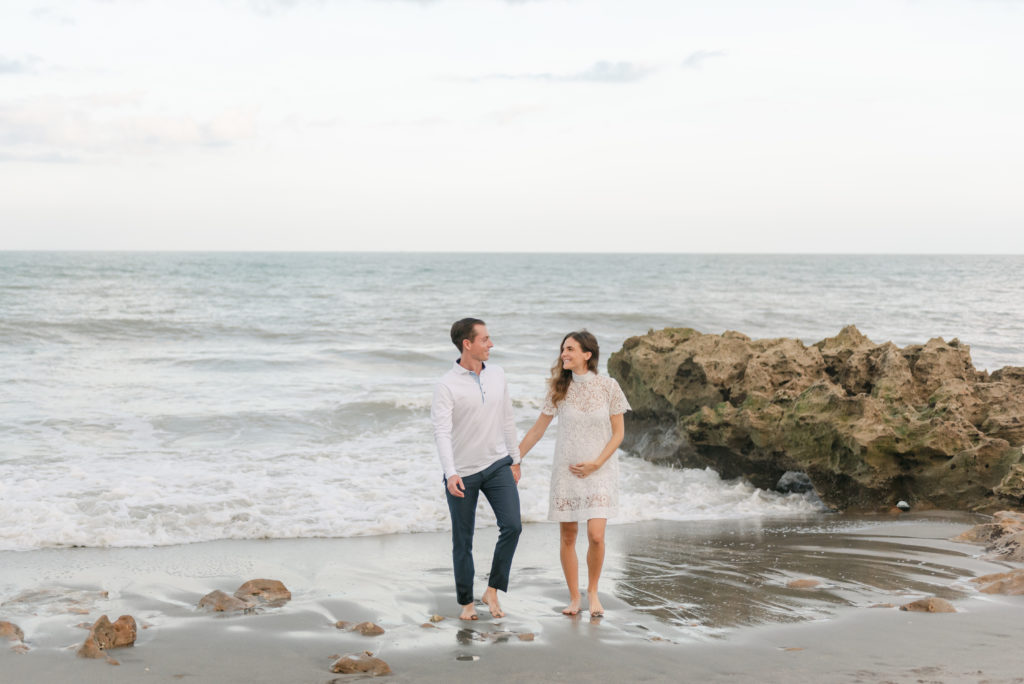 Man and woman holding hands walking away from ocean looking at each other by Jupiter photographer