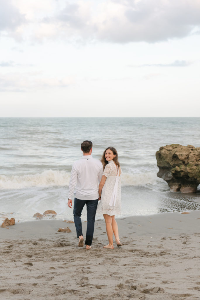 Man and woman walking towards ocean, woman looking back smiling by Jupiter photographer