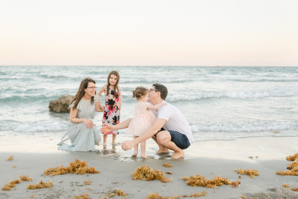 Mom and dad bending down by shoreline, each with a daughter near